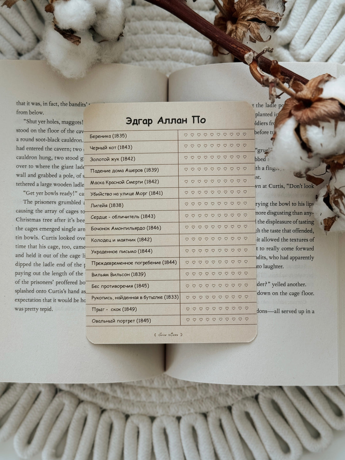 A rectangular printed card with Cyrillic text and a list of names and dates, placed on top of an open book with a dried brown flower stem across the upper right, against a light gray textured fabric background.