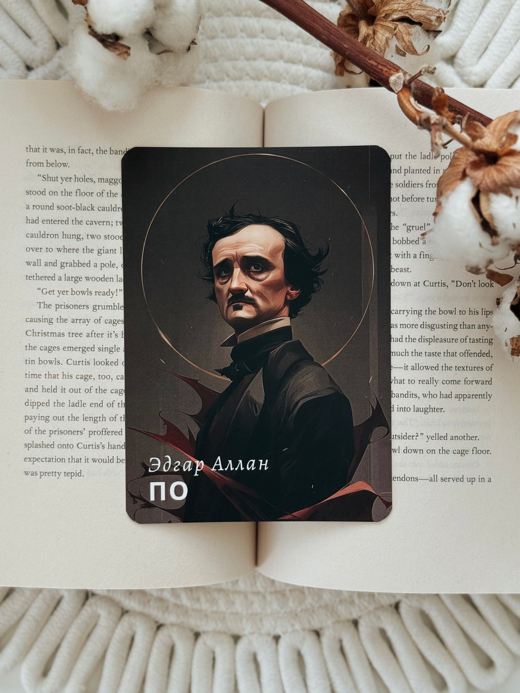 A rectangular black card featuring a portrait of Edgar Allan Poe and the text 'Эдгар Аллан ПО' in white, placed on top of an open book with printed text, styled with a wooden stick and dried flowers on a white textured blanket.