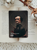 A rectangular black card featuring a portrait of Edgar Allan Poe and the text 'Эдгар Аллан ПО' in white, placed on top of an open book with printed text, styled with a wooden stick and dried flowers on a white textured blanket.