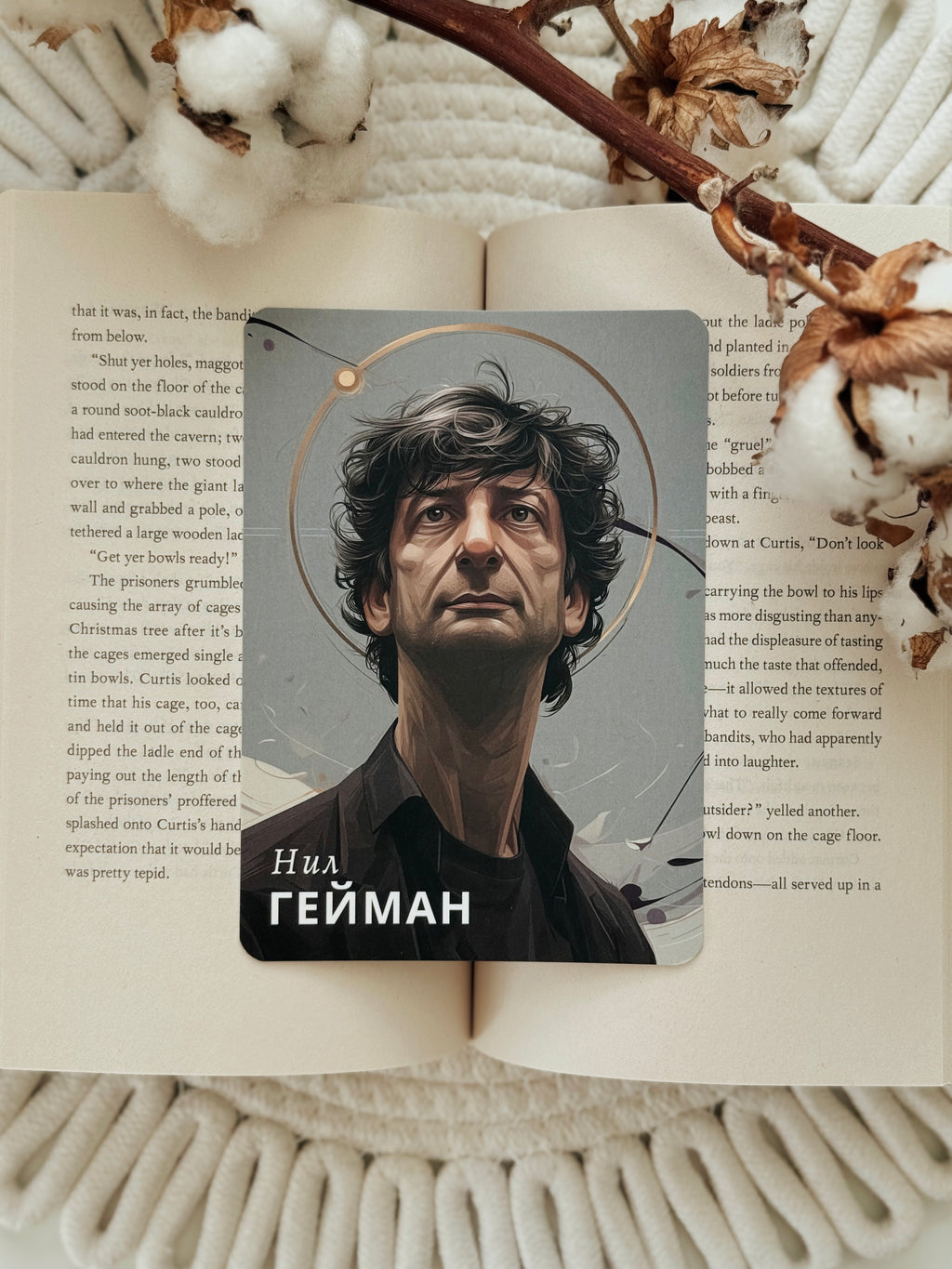 A rectangular printed card featuring a portrait of Niall Geyman with Cyrillic text at the bottom, placed on top of an open book surrounded by a knitted blanket and dried flowers.