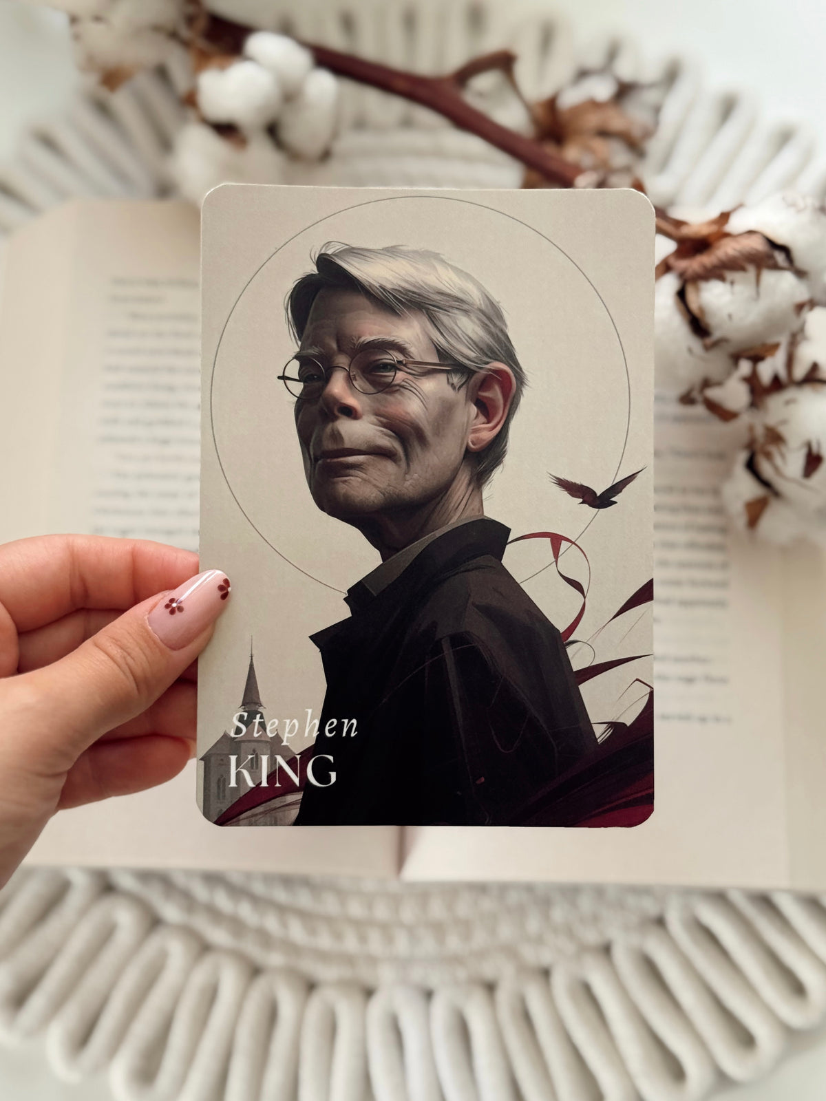 A hand with pink nail polish holds a printed card featuring an illustrated portrait of Stephen King. The card is positioned over an open book, with cotton bolls and a white woven textile in the background.
