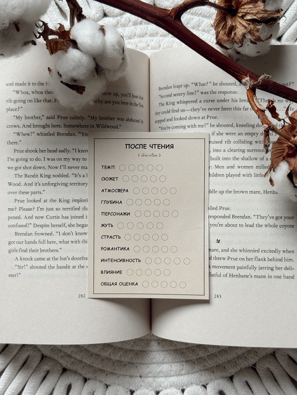 An open book spread laid on a textured white fabric background, with scattered brown leaves around it. The center page shows a printed worksheet or activity sheet in Russian with a grid of oval checkboxes.