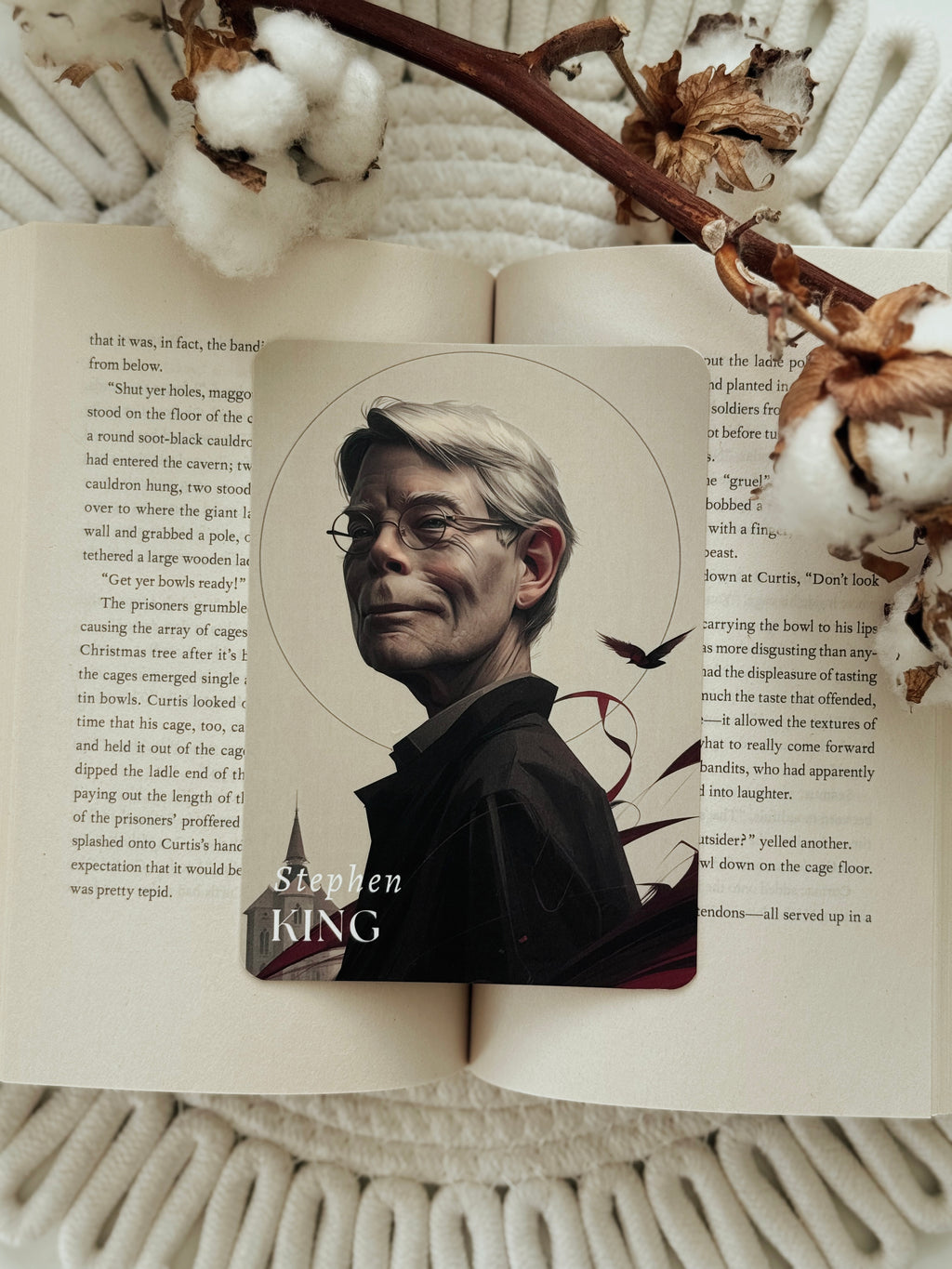 An open book laid on a white knit blanket, featuring a printed portrait of Stephen King with his name printed below. The book is styled with dried flowers and a branch on a soft white background.