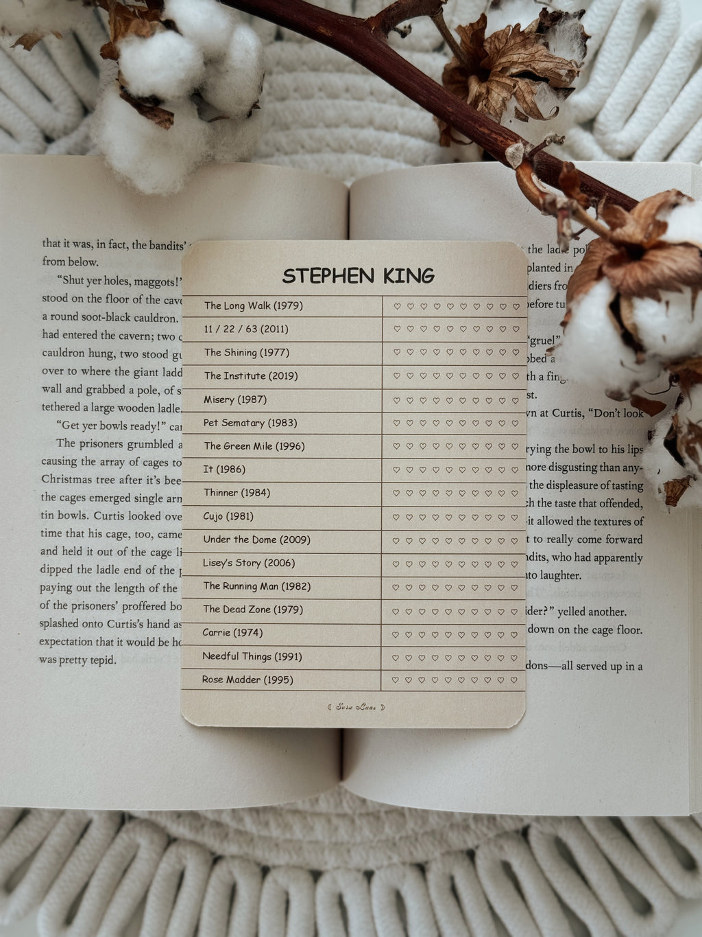 A Stephen King themed bookmark or page marker card displayed on an open book, surrounded by dried flowers on a white textured blanket.