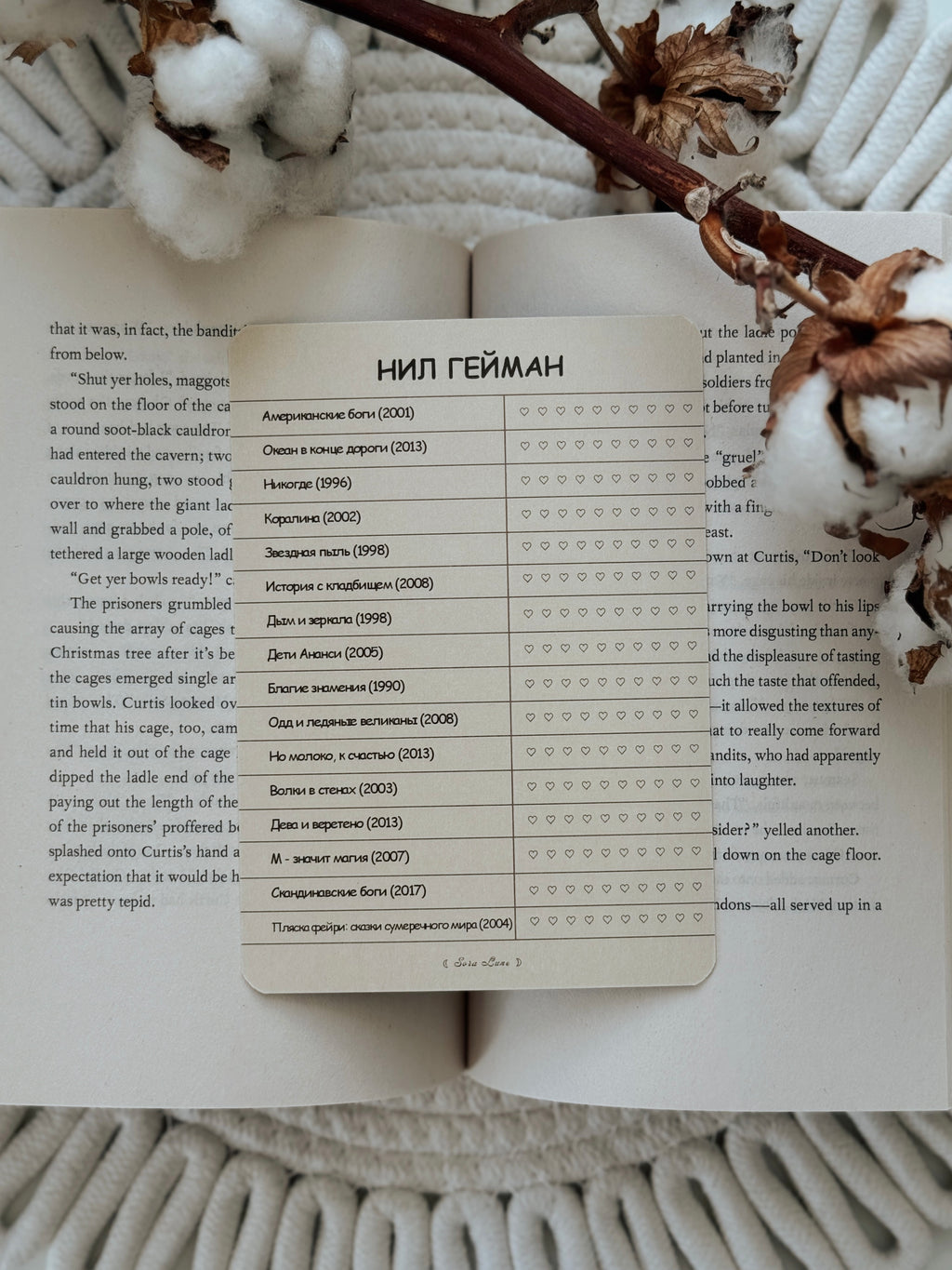 A rectangular printed card with Russian text lies on an open book, surrounded by dried brown flower petals on a white textured fabric background.