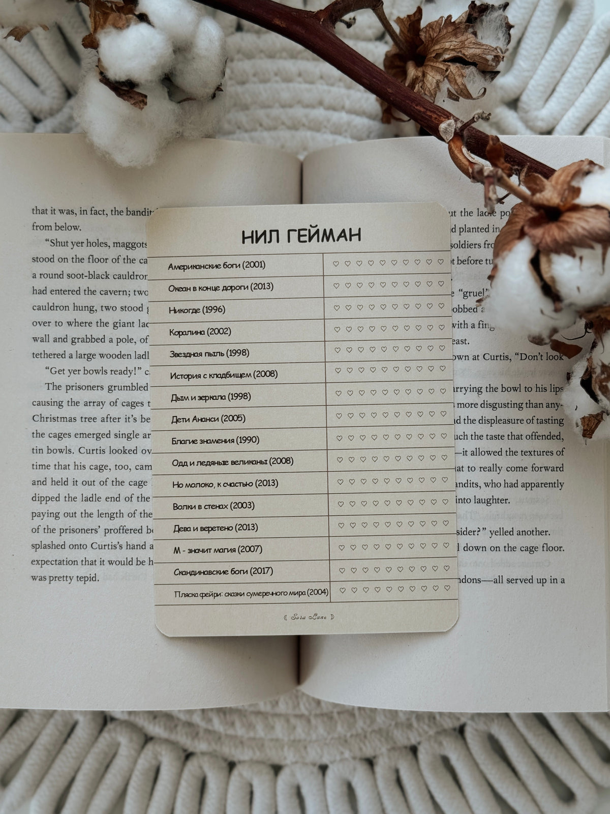 A rectangular printed card with Russian text lies on an open book, surrounded by dried brown flower petals on a white textured fabric background.