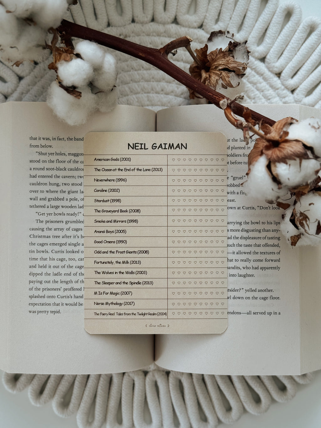 A printed book page featuring a table with the name Neil Gaiman at the top and rows of book titles and review stars, styled on an open book with dried flowers on a white textured blanket.