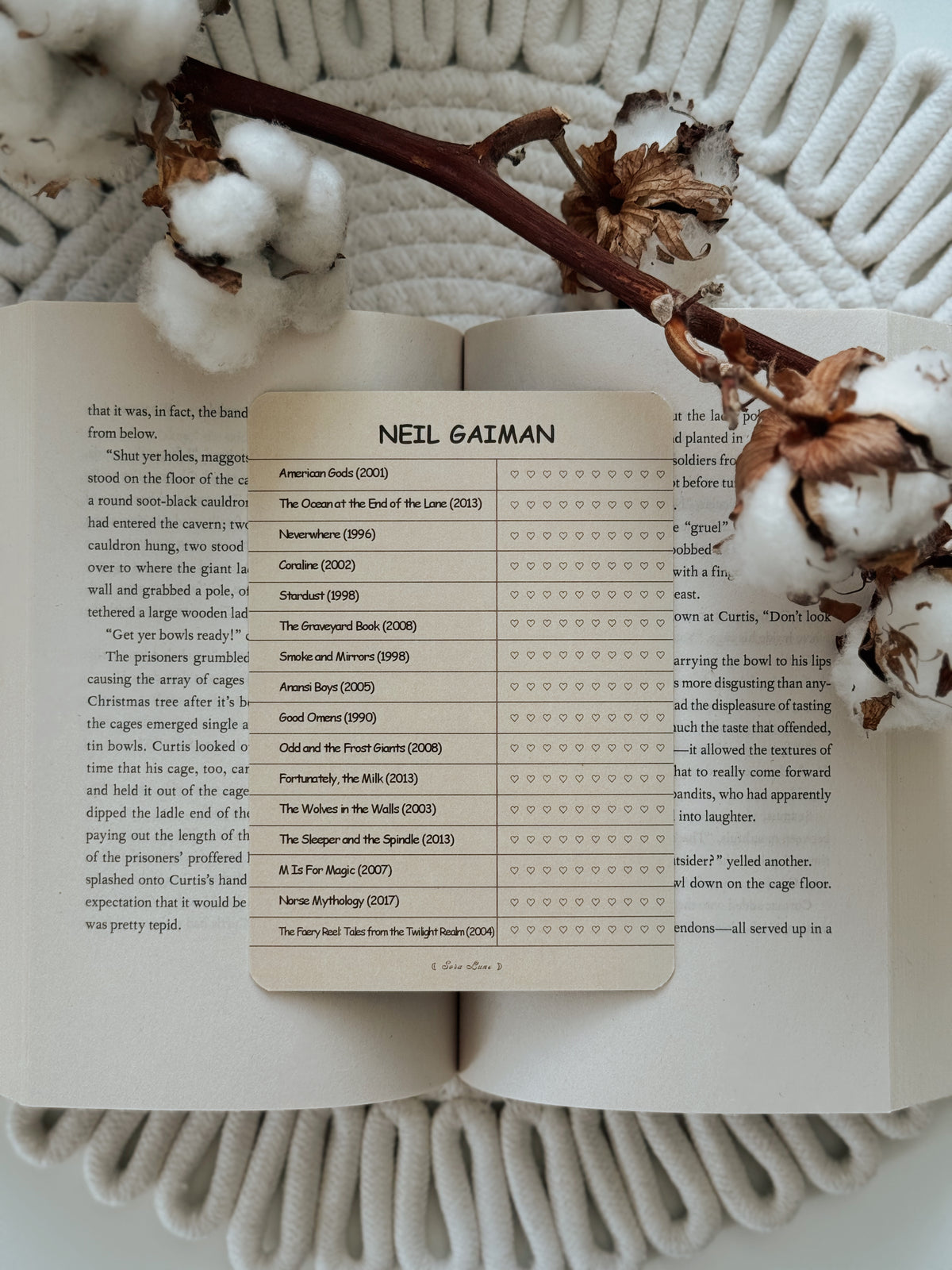 A printed book page featuring a table with the name Neil Gaiman at the top and rows of book titles and review stars, styled on an open book with dried flowers on a white textured blanket.