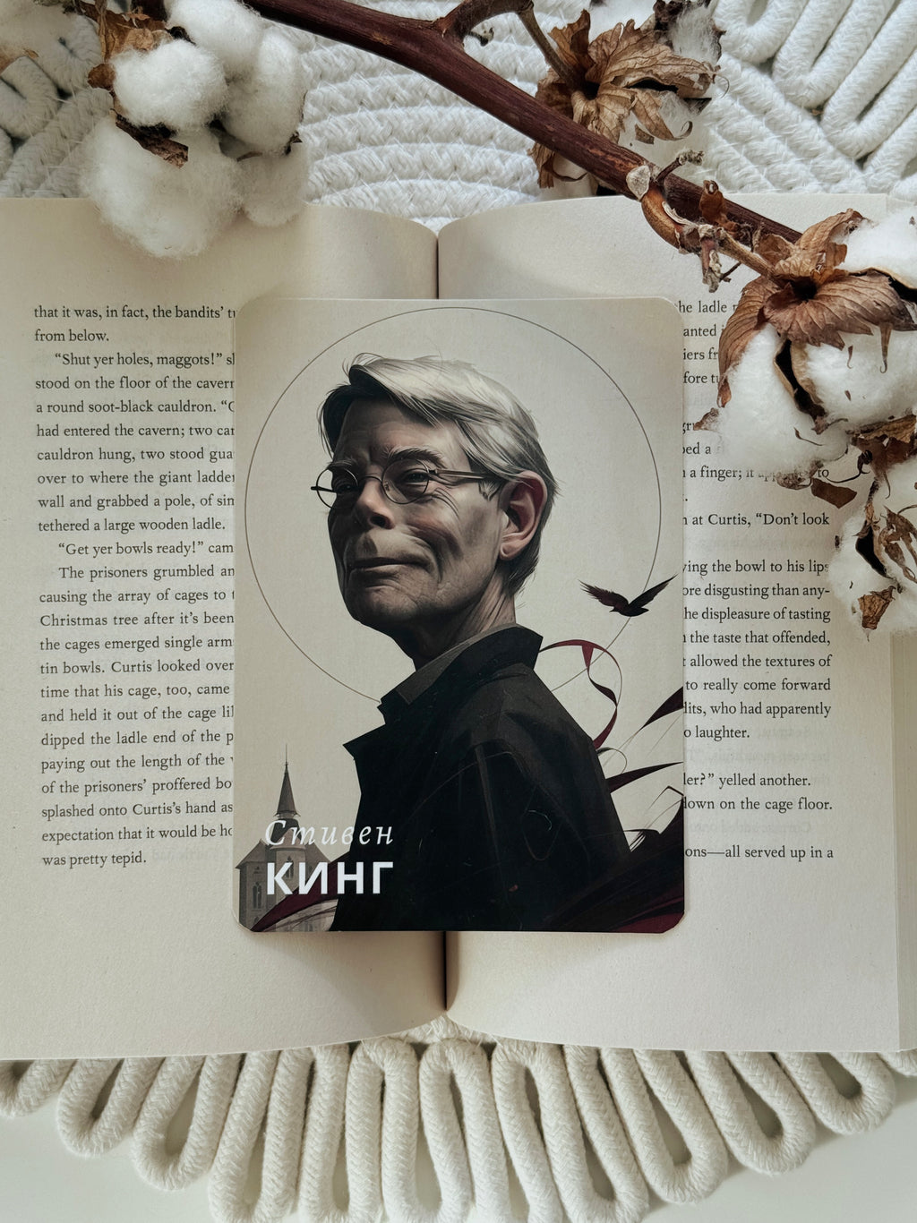 A rectangular printed card or sticker featuring a portrait of a man with glasses and the text 'Симеон КИНГ' at the bottom, placed on an open book with decorative dried leaves and white textured fabric around it.
