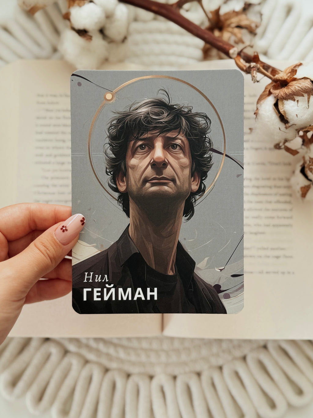 A hand holds a printed card featuring a stylized portrait of Neal Gaiman, with the text 'Neal GAIMAN' at the bottom, set against an open book and cotton stems.