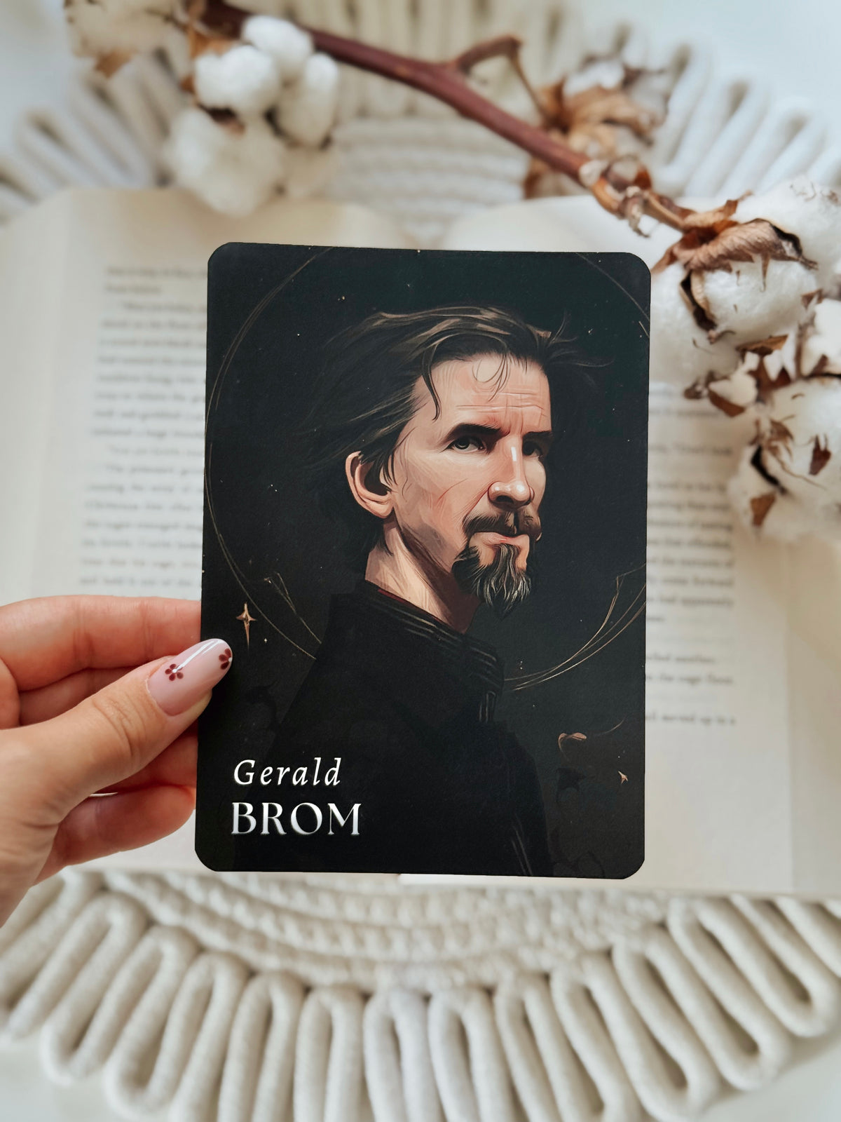 A hand holds a tarot card featuring an illustrated portrait of Gerald Brom against a dark background. The card is positioned over an open book with decorative cotton stems on a white woven mat.