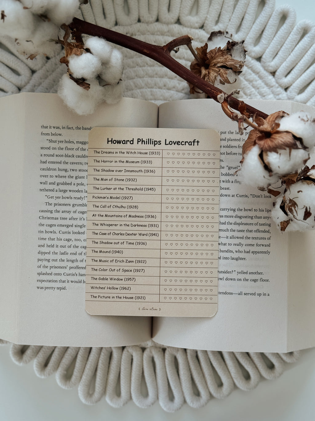 An open book spread on a white knit blanket, featuring a printed sheet titled 'Howard Phillips Lovecraft' with a list of Lovecraftiana and a star rating column. Dried botanical stems and cotton bolls are placed on top.