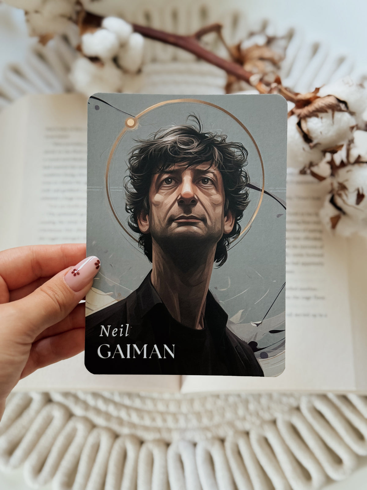 A hand holds a rectangular card featuring an illustrated portrait of Neil Gaiman against an open book with cotton stems in the background.