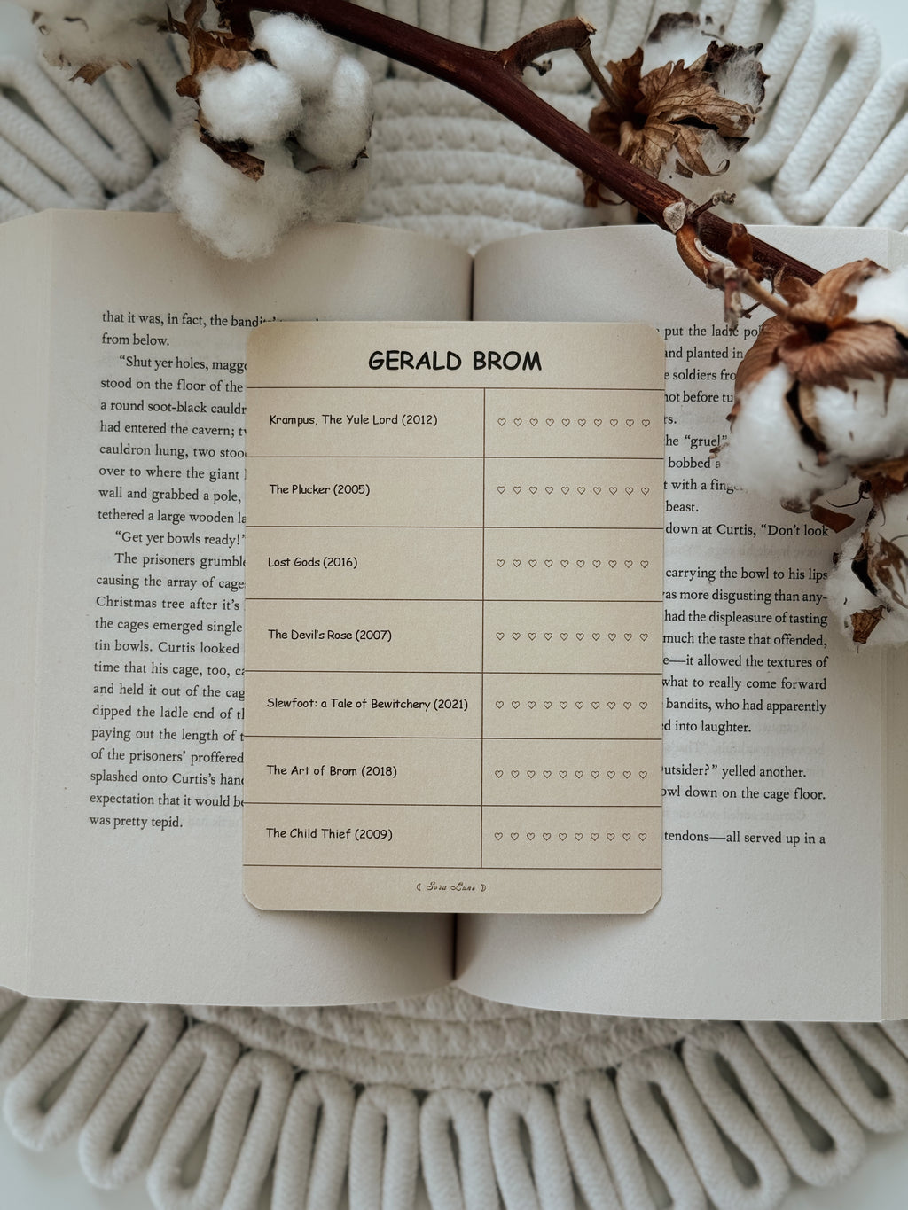A printed book page insert titled 'GERALD BROM' laid over open book pages, styled on a cream knit blanket with dried flowers and a branch around it.