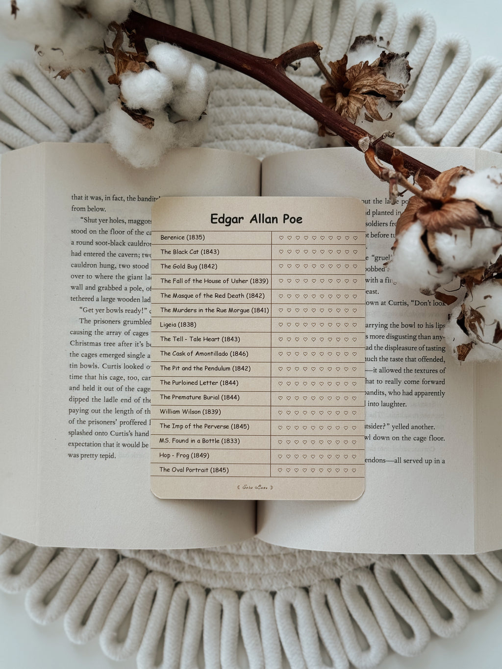 An open book spread laid on a white knit blanket, with a printed page titled 'Edgar Allan Poe' and a list of poems displayed on the right. Cotton bolls and a brown stem rest across the top of the pages.