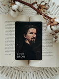 A rectangular black card featuring a portrait of Gerald Brom with the name 'Gerald BROM' printed at the bottom, placed on an open book surrounded by cotton bolls and dried floral stems.