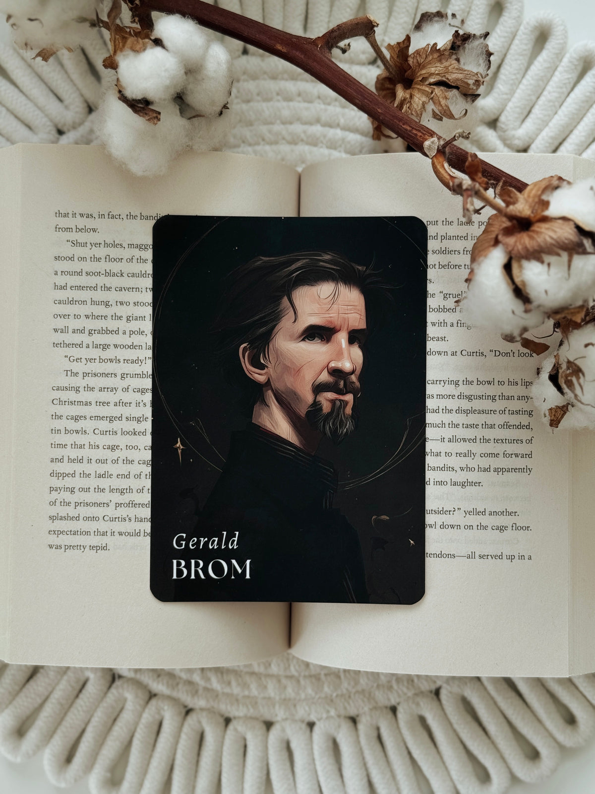 A rectangular black card featuring a portrait of Gerald Brom with the name 'Gerald BROM' printed at the bottom, placed on an open book surrounded by cotton bolls and dried floral stems.