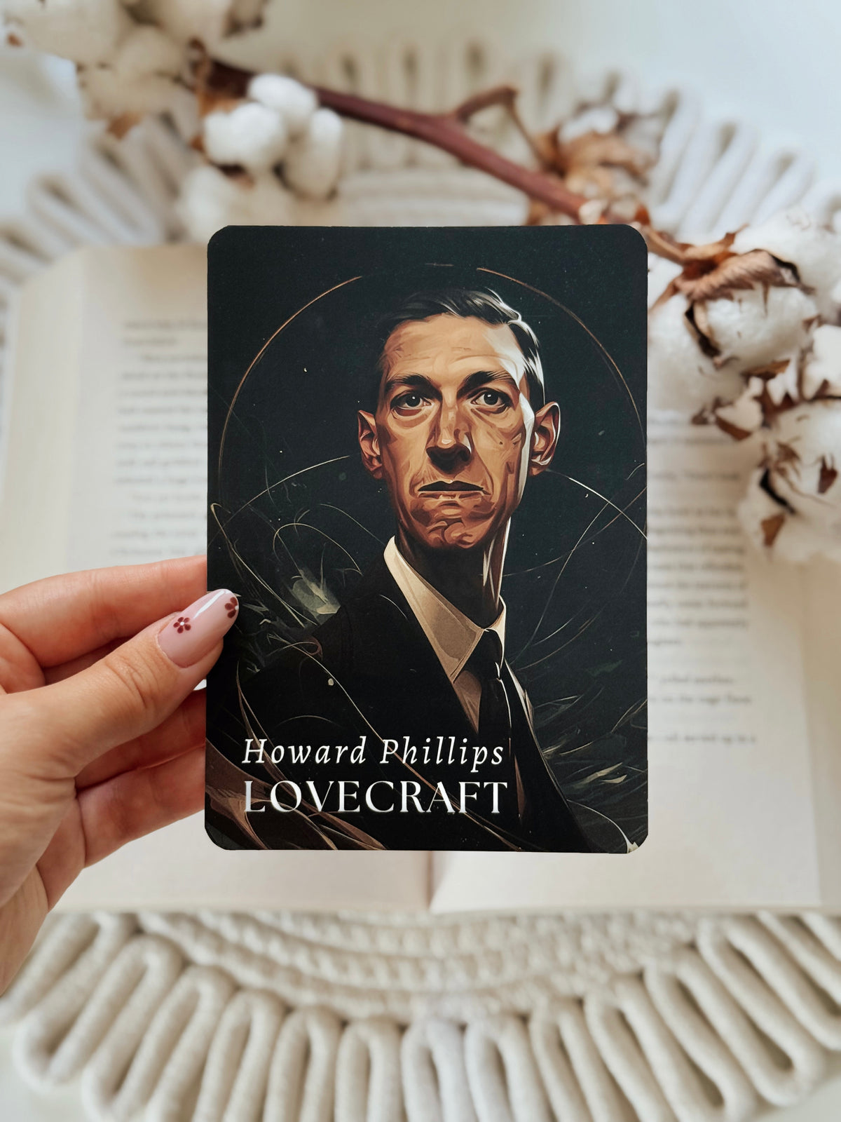 A hand holds a rectangular art print featuring a portrait of Howard Phillips Lovecraft against a dark background, with the text 'Howard Phillips LOVECRAFT' at the bottom. The scene is styled with an open book and cotton bolls in the background.
