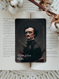 A rectangular card featuring a black portrait of Edgar Allan Poe with the text 'Edgar Allan POE' at the bottom, placed on an open book with cotton stems and a knit blanket in the background.