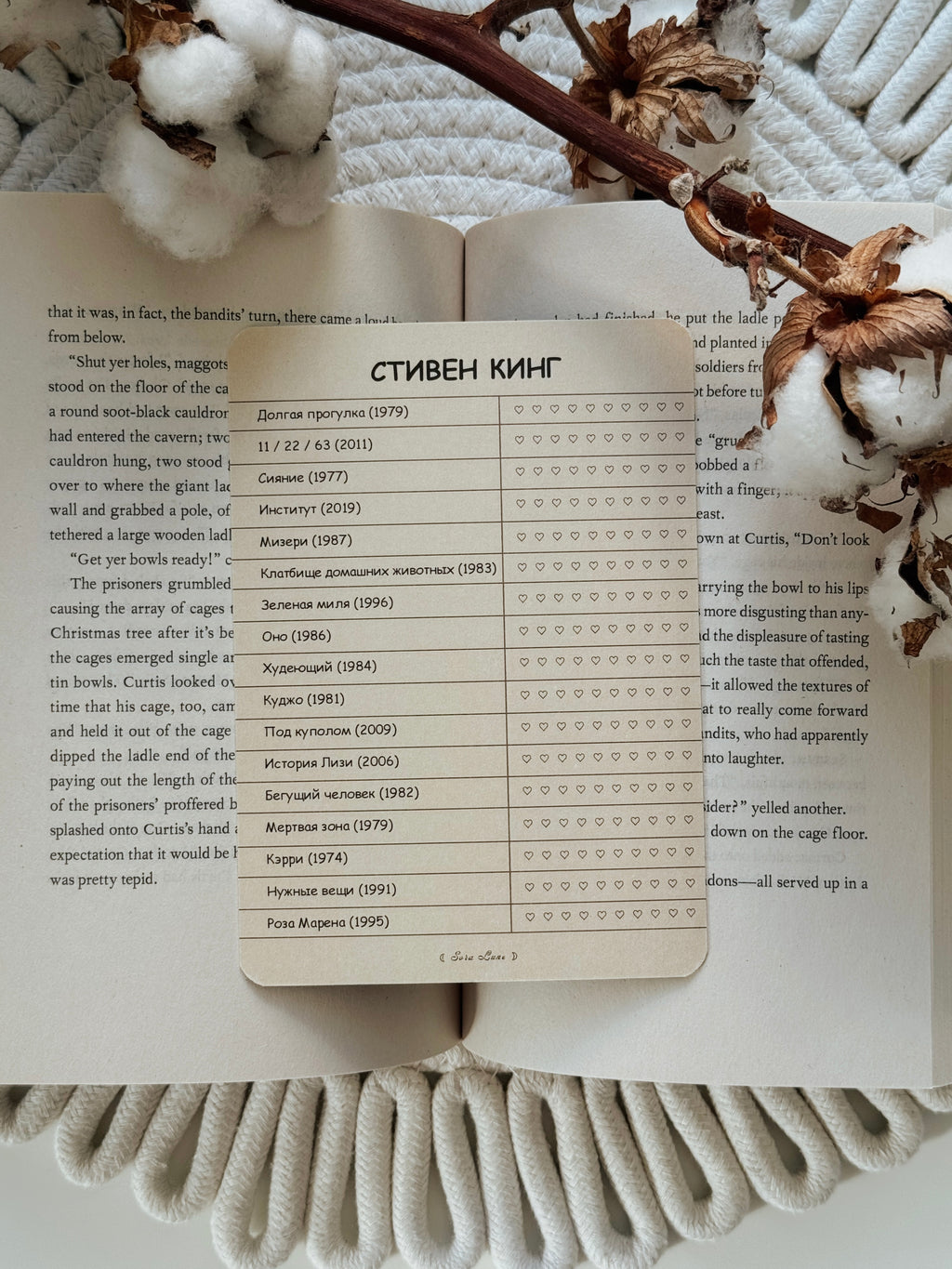 A printed worksheet titled 'STIVEN KINGS' with columns for name, year, and rating stars, laid flat on an open book with visible page text. The scene is styled with dried flowers and white cotton bolls on a soft neutral background.