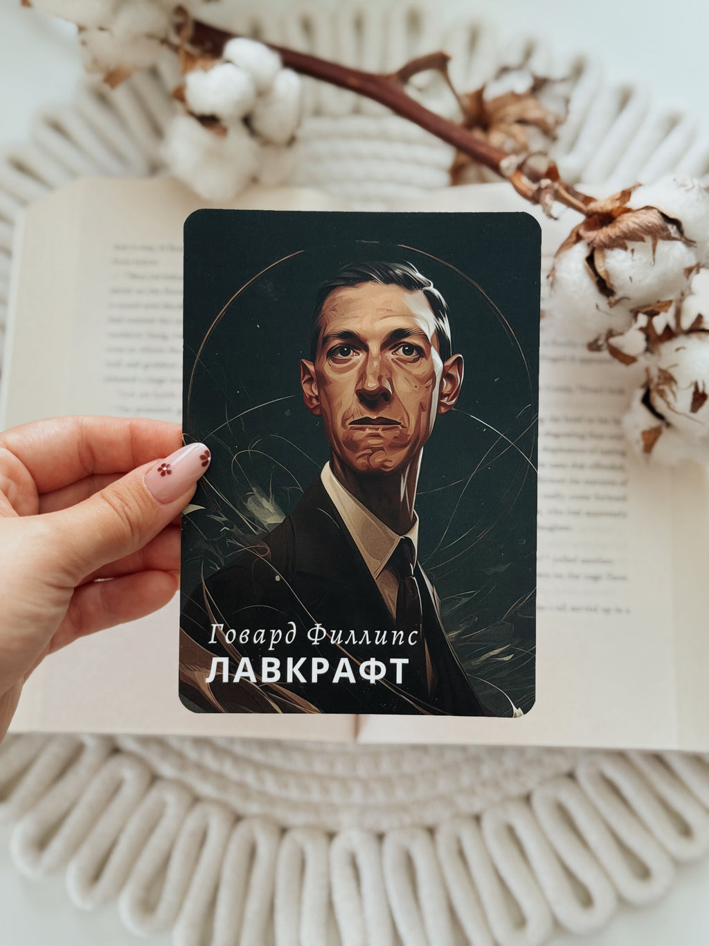 A hand holds a rectangular card featuring a dark portrait of a man in a suit with the text 'Говард Филлипс ЛАВКРАФТ' in white Cyrillic. The card is positioned over an open book on a cream-colored woven mat, with cotton stems in the background.
