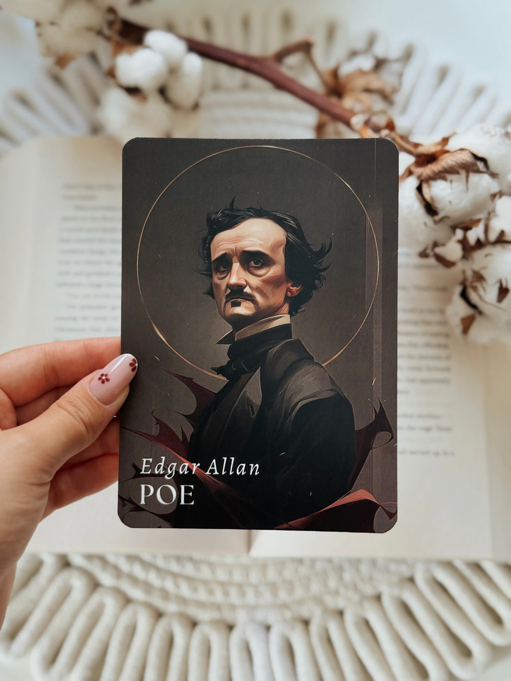 A hand holds a tarot card featuring a portrait of Edgar Allan Poe, with the text 'Edgar Allan POE' at the bottom. The background shows an open book with cotton bolls and a white woven textile.