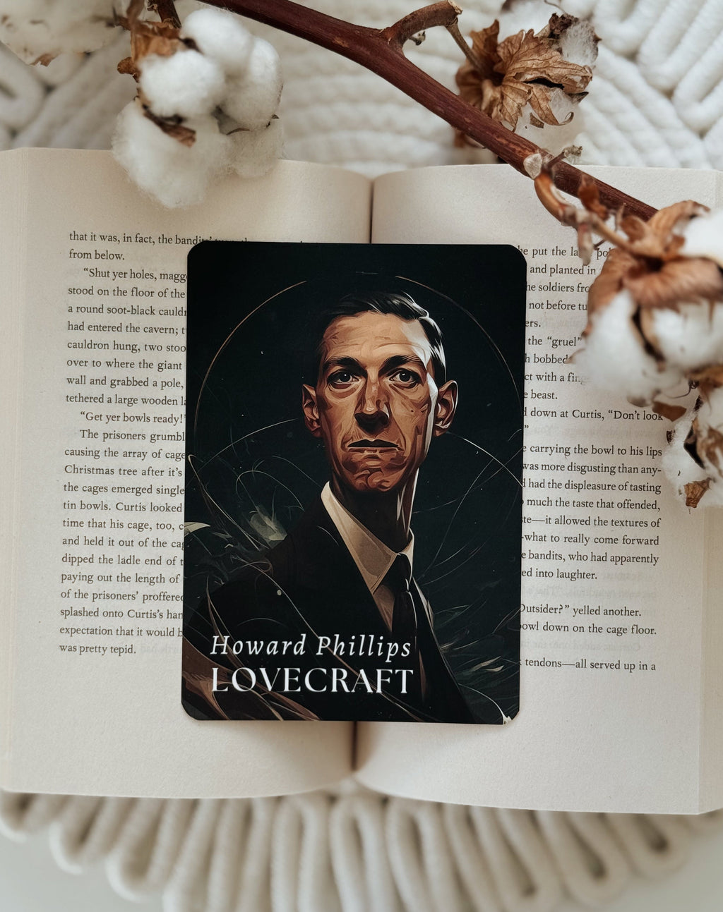 A rectangular card featuring a dark portrait of Howard Phillips Lovecraft with the text 'Howard Phillips LOVECRAFT' at the bottom, placed on an open book surrounded by cotton bolls and a dried branch.