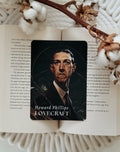 A rectangular card featuring a dark portrait of Howard Phillips Lovecraft with the text 'Howard Phillips LOVECRAFT' at the bottom, placed on an open book surrounded by cotton bolls and a dried branch.