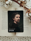 A rectangular printed card featuring a portrait of Daniel Craig as Jeremy Brock with the text 'Деникнев влетой' in white Cyrillic, placed on top of an open book with printed text, styled with a dried twig and cotton boll on a cream knit blanket.