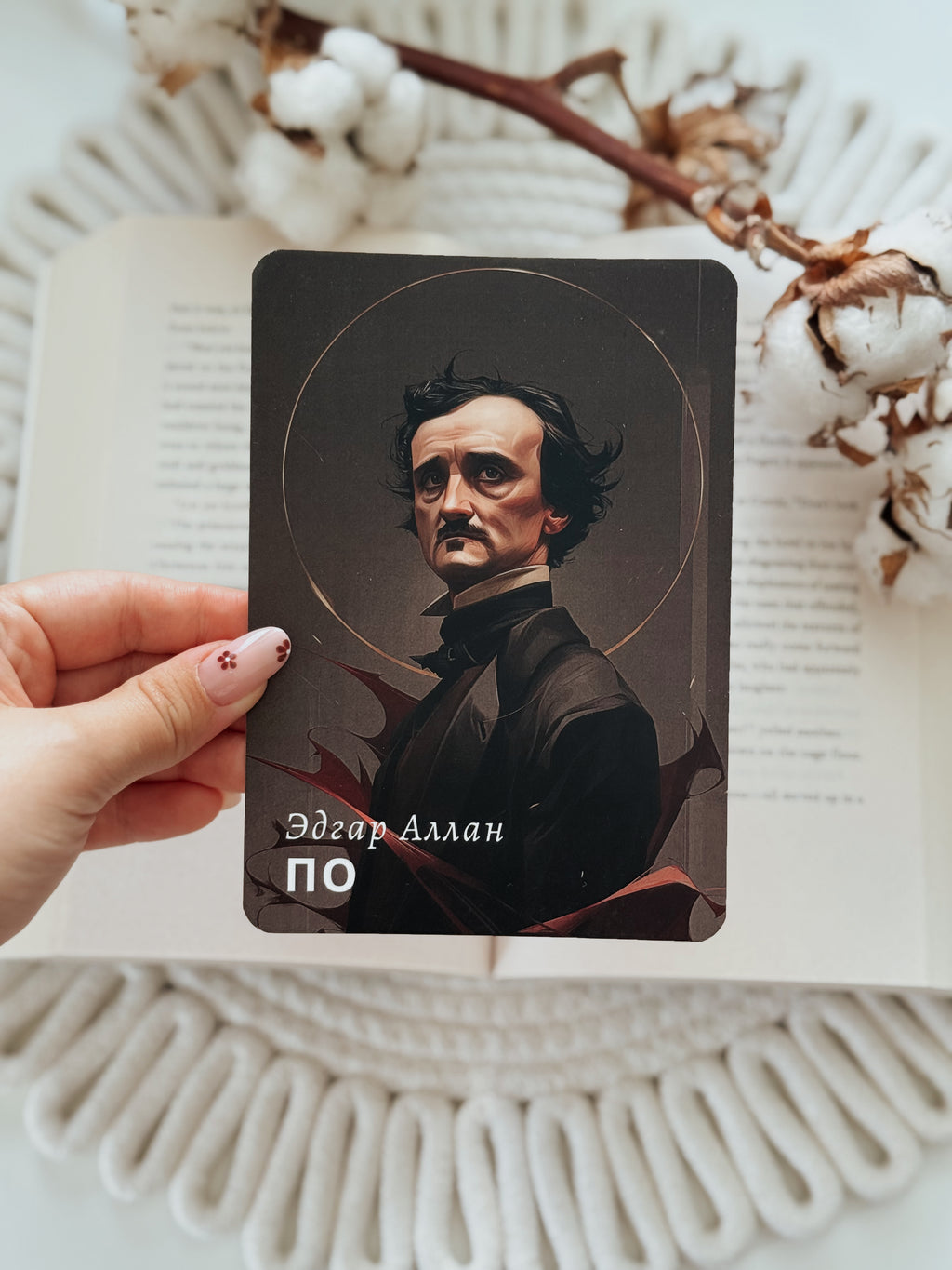 A hand holds a rectangular card featuring a portrait of Edgar Allan Poe, positioned over an open book with decorative cotton stems on a white textured surface.