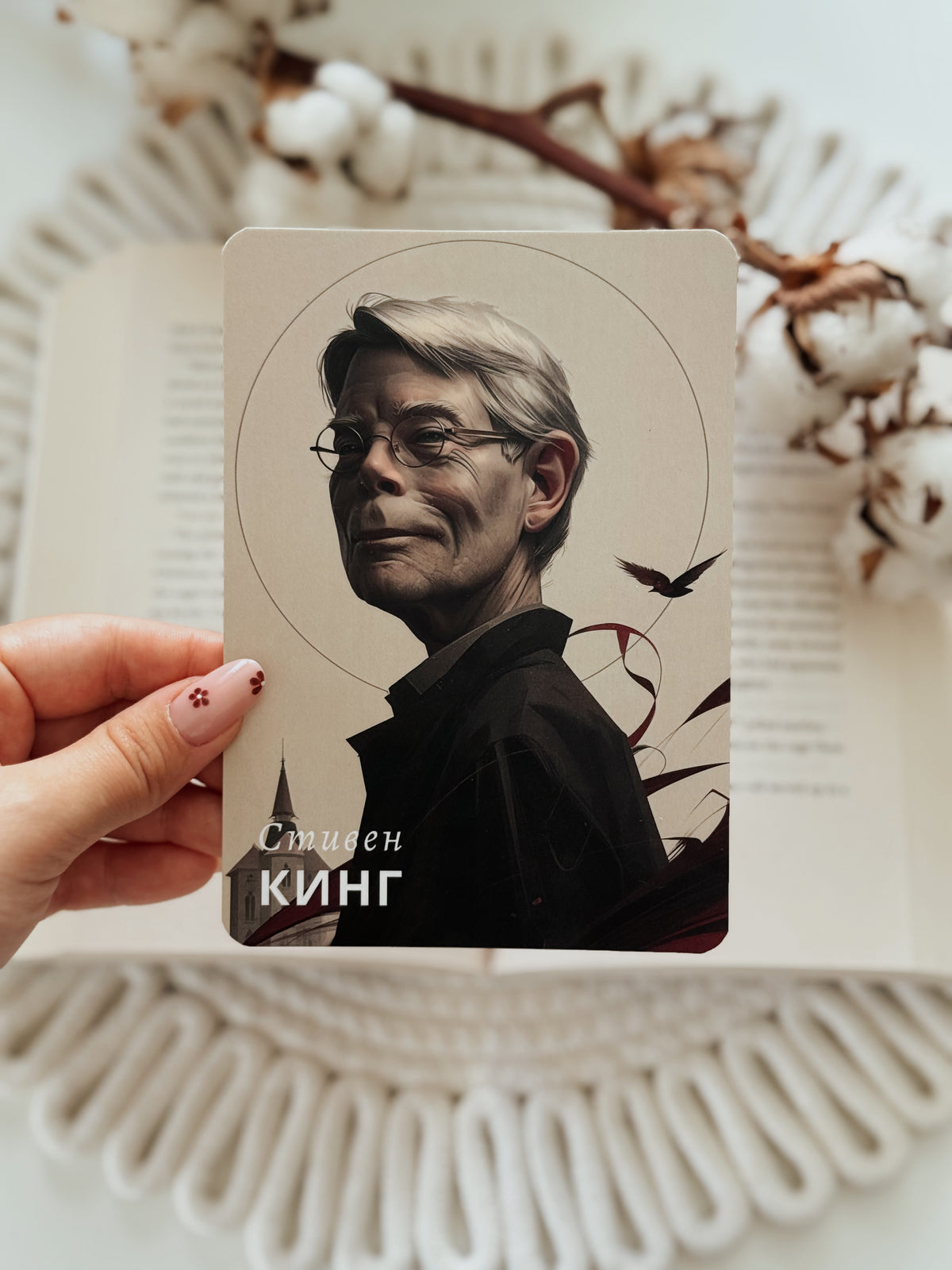 A hand holds a rectangular art print or card featuring a portrait of a man with gray hair and glasses, with the text 'Sammen Kíng' at the bottom. The background shows an open book with cotton bolls and a white woven textile.