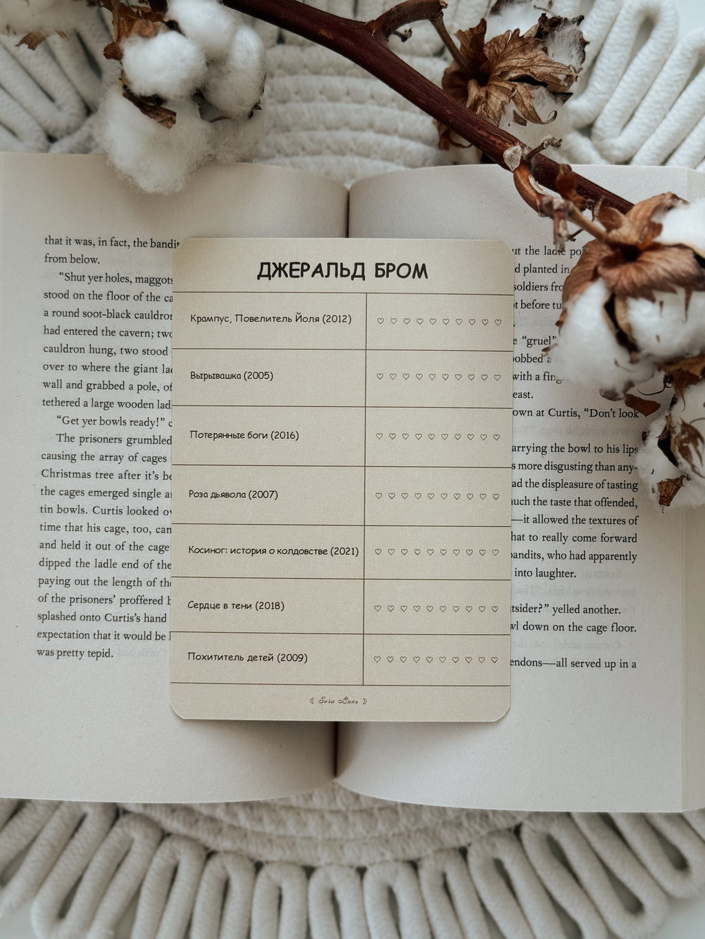 A rectangular cream-colored wooden name card with Russian text and rows of small black checkmarks, placed on an open book with dried brown flowers and white knit socks in the background.