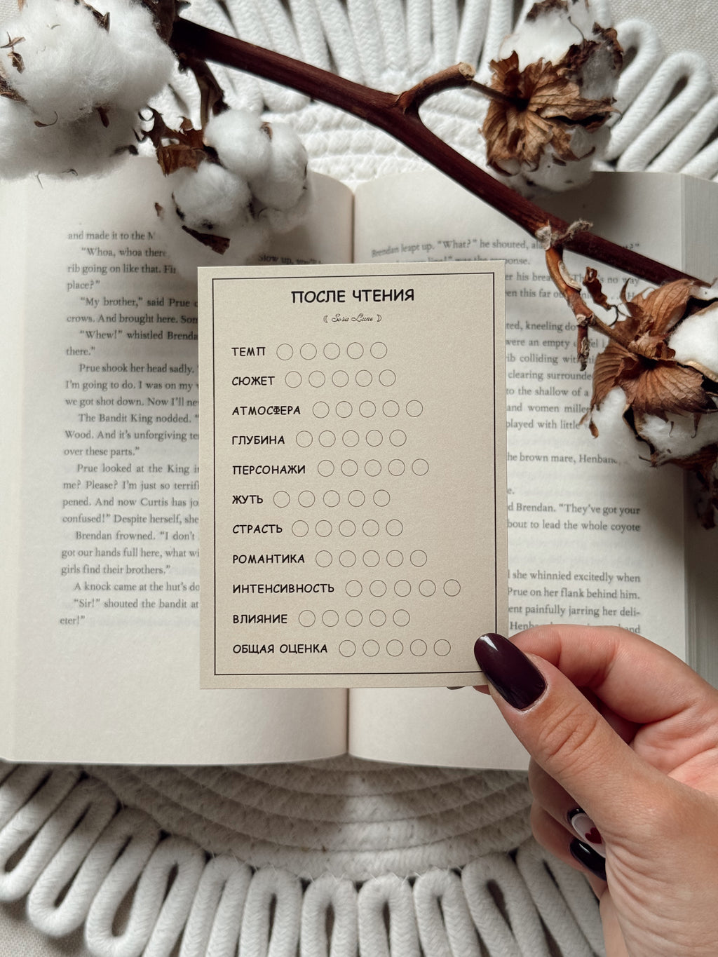 A hand with dark plum nail polish holds a Russian-language reading reflection card over an open book, styled with dried cotton bolls on a white knit blanket.